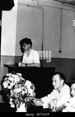 Slaviansk, 1959 anni, Energy Technical College. Un giovane laureato tiene un discorso da un podio di legno durante la difesa storica del primo diploma. In primo piano, i partecipanti seduti e i membri della commissione accademica ascoltano attentamente la presentazione del giovane specialista. Un bellissimo bouquet di margherite fresche si trova sul tavolo più vicino alla macchina fotografica. L'abbigliamento classico di metà secolo del giovane uomo cattura perfettamente l'atmosfera seria e concentrata dell'educazione tecnica sovietica. Una vita pacifica nel Donbas prima del conflitto. Foto Stock