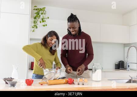 Donna indiana e uomo afroamericano che prepara la pastella all'isola della cucina con ciotola di vetro e uova Foto Stock