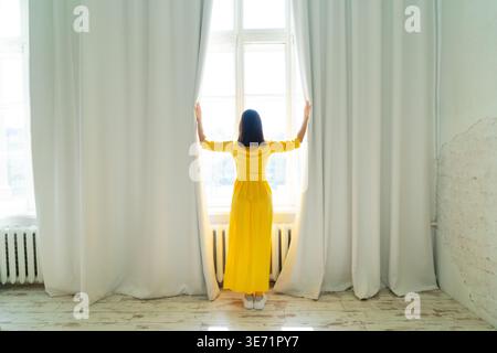 Una donna allegra vestita con un vivace abito giallo posa graziosamente e felicemente vicino alla finestra luminosa Foto Stock