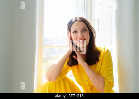 Una donna allegra vestita con un vivace abito giallo posa graziosamente e felicemente vicino alla finestra luminosa Foto Stock