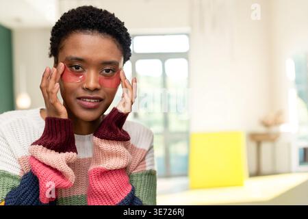 Donna afroamericana adulta che regola le toppe rosa sotto gli occhi in una stanza luminosa con un maglione grosso. Dermocosmesi, idrogel, tamponi, colorblock, cardigan, k Foto Stock