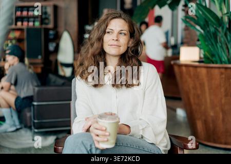 Una donna calma ed elegante siede in un caffè moderno, gustando un caldo latte. Luce naturale e vegetazione creano un'atmosfera accogliente e rilassata, perfetta per lo stile di vita, il benessere e i progetti relativi al caffè. Foto Stock