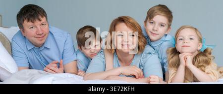 Famiglia allegra che gioca insieme a letto. I genitori trascorrono il loro tempo libero con la loro figlia e due figli. Grande famiglia caucasica che gioca a casa in camera da letto. Mamma e papà stanno ingannando con i bambini Foto Stock