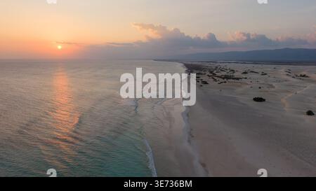 Vista aerea della tranquilla costa al tramonto con onde morbide, ampia spiaggia sabbiosa e colline lontane sotto il cielo pastello, panorama marino panoramico. Zahek Sand Foto Stock