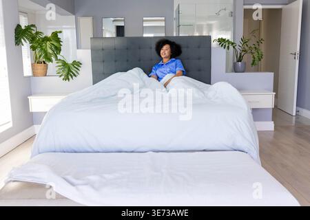 Donna di medie dimensioni seduta su un letto matrimoniale king size in camera da letto con pigiama di raso blu, piante, testata con travi a forma di ciuffo. Minimalista, moderno, luminoso, sereno, accogliente, inter Foto Stock