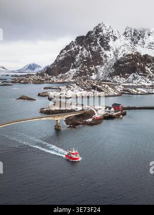 Vista aerea di una vivace barca rossa che attraversa le acque scure verso il gruppo di edifici di Henningsvær, Nordland, Norvegia. Foto Stock