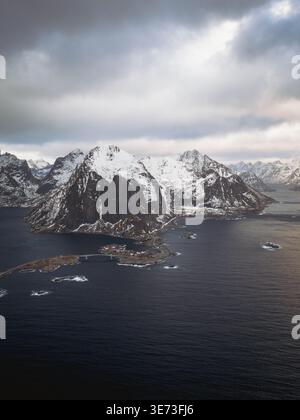 Vista aerea delle cime frastagliate innevate si innalzano bruscamente dalle acque buie e tranquille che circondano Reinebringen, Nordland, Norvegia. Foto Stock