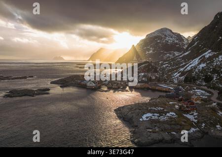 Vista aerea della luce del sole dorata che penetra attraverso le spettacolari nuvole sulle isole Lofoten, proiettando un caldo bagliore sulle montagne innevate e sul tra Foto Stock