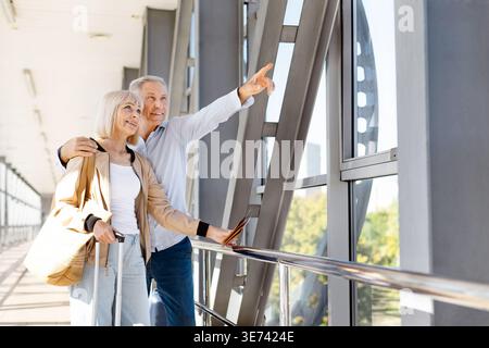 Uomo maturo che punta attraverso la finestra dell'aeroporto mentre abbraccia la moglie con i bagagli Foto Stock