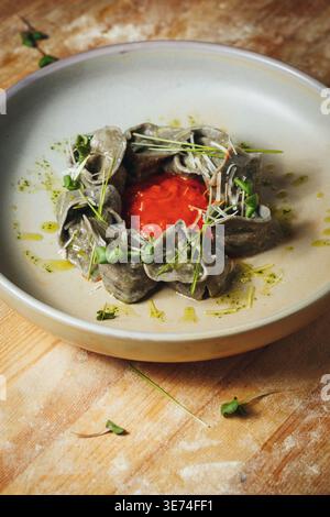 Tortellini gourmet a inchiostro nero serviti con vivace salsa di pomodoro microgreen freschi e scaglie di parmigiano su un piatto di ceramica. Foto Stock