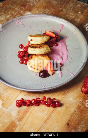 Tradizionali frittelle di formaggio fresco con sciroppi dorati serviti con fragole fresche ribes rosso e una vivace salsa di bacche rosa su un piatto di ceramica grigia. Foto Stock