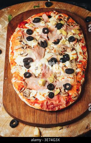 Tradizionale pinsa romana capricciosa italiana condita con salsa di pomodoro mozzarella prosciutto olive nere carciofi e funghi su una tavola di legno. Foto Stock