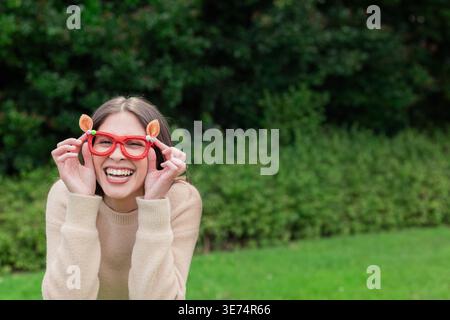 Donna pendente della metà degli anni '20 con occhiali rossi con orecchie arancioni, maglione beige nel parco, spazio fotocopie. All'aperto, risate, siepi, prato, maglia, giardino, nat Foto Stock
