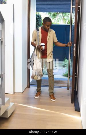 Uomo afroamericano che entra a casa alla soglia, portando con sé una borsa a rete con prodotti, indossando uno zaino. Ingresso, borsa tote, alimentari, luce del giorno, pendolari, esterno Foto Stock