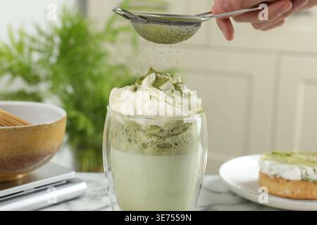 Donna setacciando polvere di matcha su bevanda a tavola, primo piano Foto Stock
