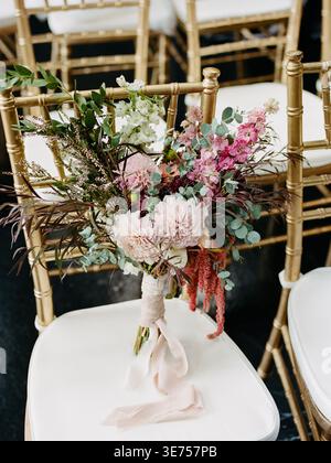 Splendido bouquet nuziale collocato su una sedia in un luogo allestito per una cerimonia con sedie dorate e sedie bianche Foto Stock