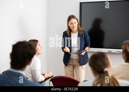 Donna di mezza età che si trova vicino a una lavagna interattiva e che comunica con studenti adulti durante una lezione di lingua straniera Foto Stock