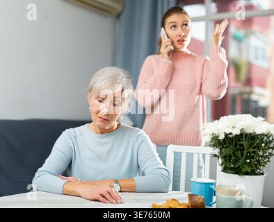 Donna anziana turbata seduta a tavola mentre figlia adulta irritata che parla al telefono Foto Stock