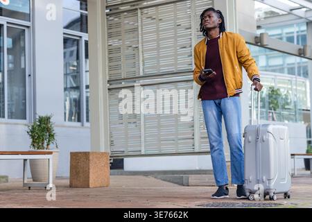 Uomo afroamericano in giacca di senape in piedi a controllare il telefono che tiene la valigia d'argento, copia spazio. Viaggiatore, bagagli, dispositivo, all'aperto, urbano, plaza, Foto Stock