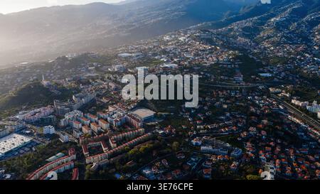 Vista aerea di Funchal, Madeira, con edifici bianchi, tetti in terracotta, strade tortuose, viadotto della circonvallazione e chiesa in cima a una collina al mattino o in ritardo a poppa Foto Stock