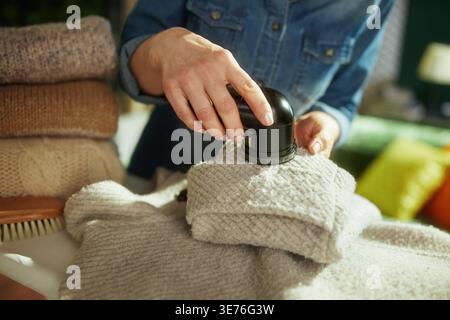Una persona con una camicia in denim utilizza con cura un rasoio per tessuti su un cappotto invernale ripiegato per garantire una qualità duratura durante la transizione stagionale al guardaroba. Foto Stock