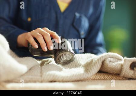 Una persona con una camicia blu in denim aziona con attenzione un rasoio per tessuti nero su un cappotto invernale beige leggero per rimuovere pelucchi e pilling, garantendo un tessuto più lungo Foto Stock