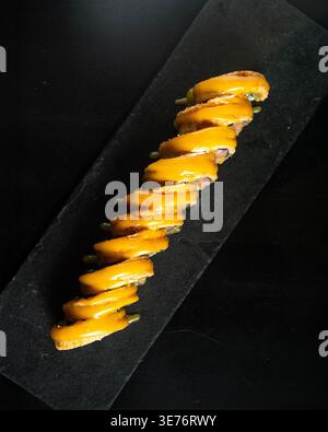 I rotoli dorati poggiano sulla pietra nera. Ogni fetta mostra un riempimento fresco e vivace. Il sushi e' ordinato per l'eleganza. Lo sfondo scuro migliora i colori. Per Foto Stock