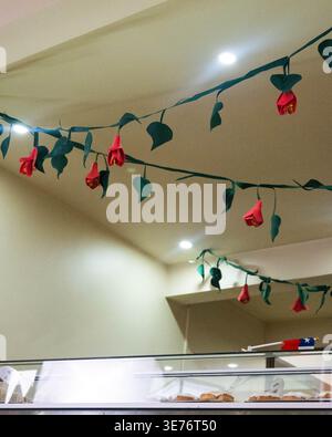 Le rose di carta si sgretolano dalle stringhe del soffitto. Le foglie verdi completano i fiori rossi. Le luci illuminano delicatamente le decorazioni. Espositore per prodotti da forno Foto Stock