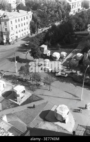 Sloviansk, 1997, via Yunykh Kommunarov (ora via Bankovskaya). Una ripida foto in bianco e nero rialzata che guarda direttamente in basso verso un vivace incrocio cittadino. La cornice cattura una vivace piazza pedonale piena di distinti chioschi commerciali esagonali, circondati da auto in movimento e dalla gente del posto. Delimitato da un classico edificio storico d'angolo e da un alto blocco residenziale, questa autentica immagine d'archivio offre un affascinante scorcio sul commercio a livello di strada in trasformazione in un tranquillo Donbas. Foto Stock