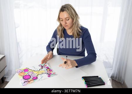 Una donna siede a un tavolo in una stanza ben illuminata, colorando con un disegno floreale su carta. Si concentra su un progetto artistico che promuove la creatività e il benessere mentale. Foto Stock