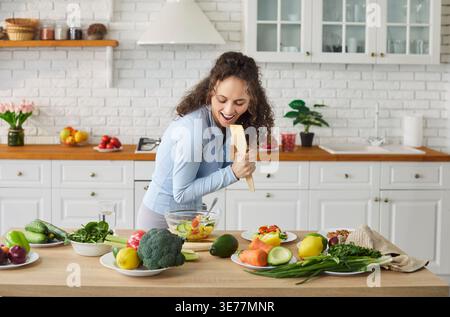 Donna che canta giocosamente nella spatola in piedi in cucina moderna, frutta e verdura fresche Foto Stock