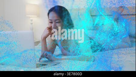 Donna coreana sdraiata che indossa tanktop rosa, sorride al laptop in camera da letto, rivestimento blu Foto Stock