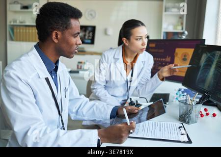 Medico afroamericano maschile e medico asiatico donna che esamina la radiografia toracica e i dati dei pazienti sui monitor. Utile per lavoro di squadra, diagnosi, radiologia, marketing medico Foto Stock
