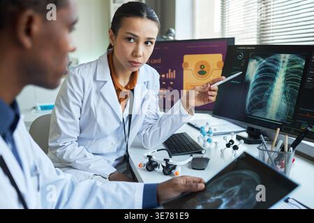 Ritratto di una donna asiatica che discute la scansione polmonare con un collega maschio nero, rivedendo l'imaging diagnostico sui monitor. Ideale per il lavoro di squadra, la radiologia e il marketing diagnostico nel settore sanitario Foto Stock