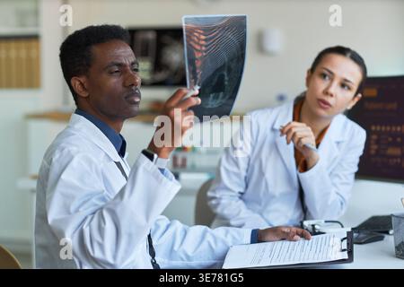 Shot medio di un medico nero che esamina la radiografia mentre una collega si consulta in clinica, supportando la diagnosi e il lavoro di squadra. Ideale per marketing sanitario, radiologia, formazione medica Foto Stock