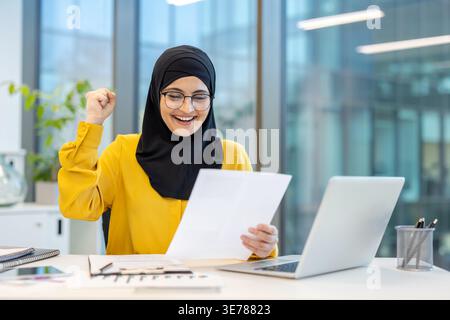 Donna d'affari musulmana che indossa hijab e occhiali che tira con il pugno alzato, celebra il successo professionale e legge buone notizie sul business ufficiale Foto Stock