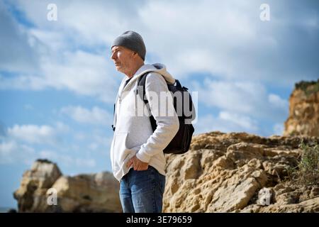 uomo che indossa un cappello di lana e una felpa con cappuccio in piedi sulle rocce con uno zaino, che guarda al mare Foto Stock