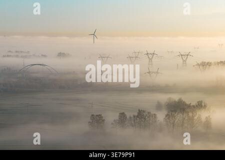 Vista aerea di un paesaggio avvolto da una nebbia argentea, un ponte e linee elettriche emergono dalla nebbia, con una sola turbina eolica alta, Wiślina, P. Foto Stock