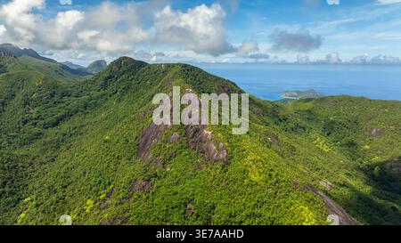 Una montagna verdeggiante coperta da fitta vegetazione, che si innalza bruscamente sotto un cielo azzurro limpido con alcune nuvole. Seychelles, Mahe. Foto Stock