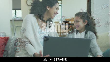 Madre e figlia sorridenti che condividono il laptop sul divano di casa, indossando cardigan con sovrapposizione di rete Foto Stock