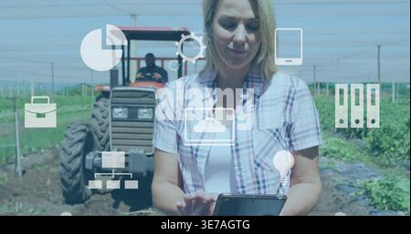Tenendo una donna che indossa una camicia a quadri con un tablet sul campo, mostrando le icone digitali e il trattore dietro di sé Foto Stock