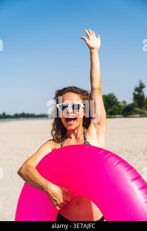 Donna allegra sulla spiaggia con anello gonfiabile rosa che ondeggia la mano, atmosfera di vacanza. Foto Stock