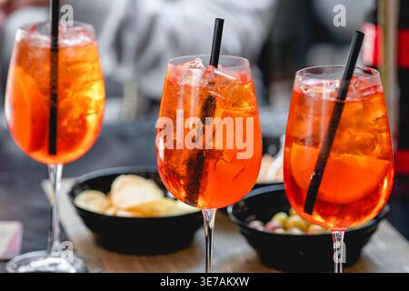 Vivaci cocktail Spritz arancioni con fette di ghiaccio e agrumi serviti in bicchieri da vino con cannucce, accompagnati da spuntini in un ambiente informale Foto Stock