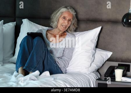 Donna anziana seduta sul letto con un tablet in camera d'albergo Foto Stock