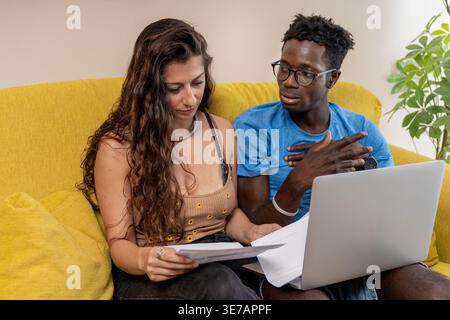 La coppia di maschi neri e caucasici collabora alle loro finanze di casa rivedendo documenti con un laptop seduto sul divano di casa Foto Stock