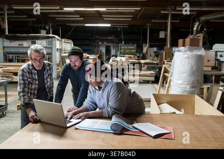 Donna nera in piedi con carpentieri caucasici che utilizza un computer portatile in un negozio di lavorazione del legno Foto Stock