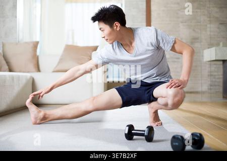 Uomo cinese che fa esercizi di stretching e guarda lontano mentre accovacciato in salotto vicino a manubri a casa Foto Stock