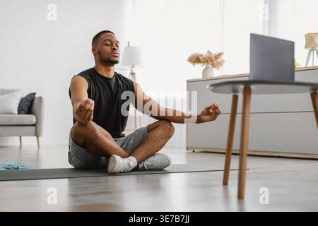 Uomo nero a occhi chiusi che medita in posizione di loto sul tappetino da yoga vicino al tavolo con il portatile nel soggiorno di casa Foto Stock