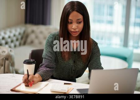 Donna nera che scrive su un notebook che guarda il portatile sul tavolo in salotto Foto Stock