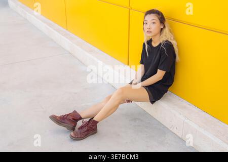 Donna cinese seria seduta con le mani tra le gambe appoggiate sul muro giallo Foto Stock
